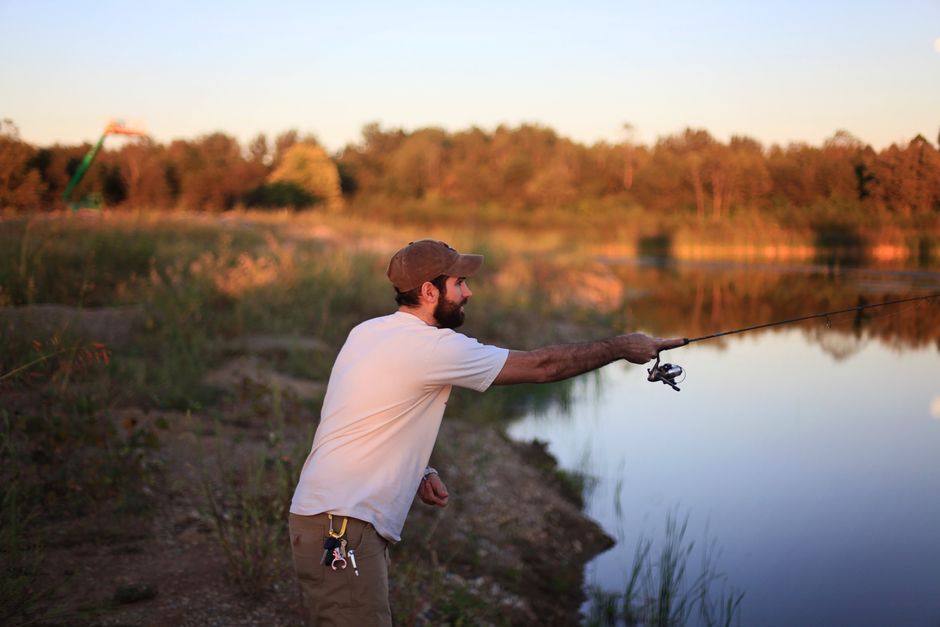 A man in casual attire fishes by a calm lake, reflecting a scenic sunset outdoors.